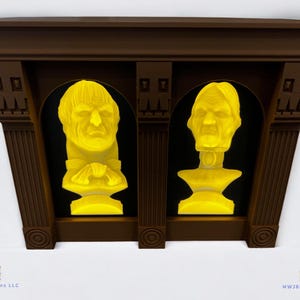 May include: A brown wooden frame with two yellow illuminated busts of people inside. The frame is mounted on a white wall.