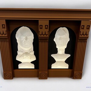 May include: A brown wooden frame with two white busts of people inside. The busts are set against a black background. The frame has a decorative top and columns on the sides.