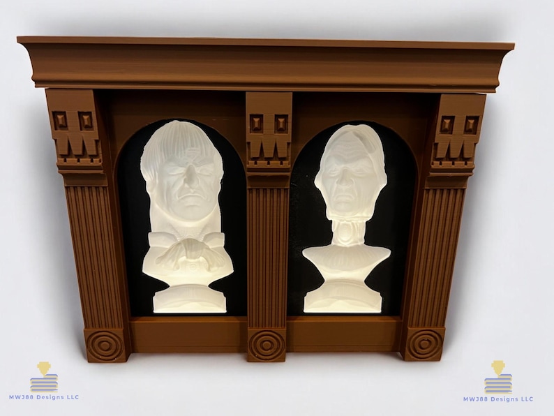 May include: A brown wooden frame with two white busts of people inside. The busts are set against a black background. The frame has a decorative top and columns on the sides.