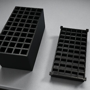 Puede incluir: Dos organizadores negros con patrón de rejilla. Uno es rectangular y vertical, mientras que el otro es más pequeño y rectangular. Los organizadores están hechos de un material sólido y están diseñados para guardar objetos pequeños. El fondo es una superficie gris claro.