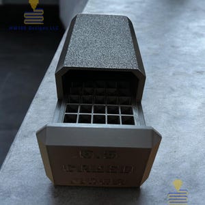 Könnte beinhalten: Eine graue, offene Box mit strukturiertem Deckel und gitterartigem Innenraum. Auf der Vorderseite der Box ist der Text "US CREED INJECTOR" eingeprägt. Die Box besteht aus mattem Material und steht auf einer grauen Oberfläche.