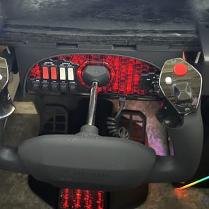 May include: A black flight simulator control panel featuring a yoke, control sticks, and various switches. The panel has red backlighting and is mounted under a dark surface. The yoke is grey and has a silver shaft.