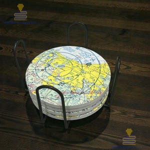 May include: A set of circular coasters with a map design, held in a black metal holder. The coasters feature a yellow, blue, and white aviation chart design. The holder has a simple, functional design.