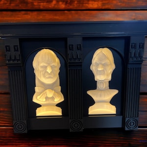 May include: Two white plaster busts of people with a black frame around them. The busts are lit from behind, creating a halo effect around the heads.