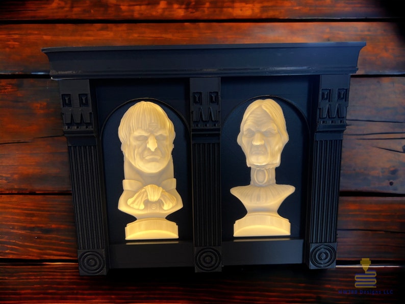 May include: Two white plaster busts of people with a black frame around them. The busts are lit from behind, creating a halo effect around the heads.
