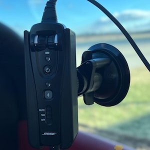 Pode incluir: Um dispositivo de áudio Bose preto com um suporte de ventosa, com um botão de ligar/desligar, um símbolo Bluetooth e um controle deslizante para silenciar, misturar e desligar. O dispositivo está montado em um vidro de carro, com o texto "MWJ88 Designs LLC" visível.