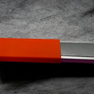 Könnte beinhalten: Nahaufnahme eines rechteckigen, silberfarbenen Objekts mit einer orangefarbenen Kappe. Das Objekt befindet sich auf einer grauen Oberfläche. Die orangefarbene Kappe befindet sich auf der linken Seite des Objekts.