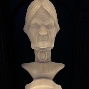 May include: A white bust sculpture of a woman with a stern expression. The sculpture is detailed with a textured surface and a decorative collar.