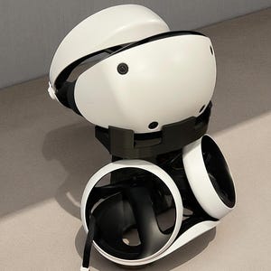 Puede incluir: Un casco y unos mandos de realidad virtual blancos y negros se muestran sobre un soporte negro. El casco tiene un diseño curvo con detalles en negro. Los mandos tienen un diseño circular con interiores negros y exteriores blancos. Una correa negra cuelga de un mando.
