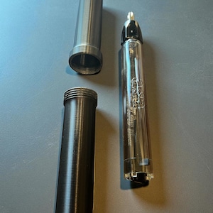 May include: An exploded view of a pen with a clear barrel and a black cap. The pen is disassembled, with the barrel, cap, and a cylindrical metal sleeve separated. The pen is on a gray surface.