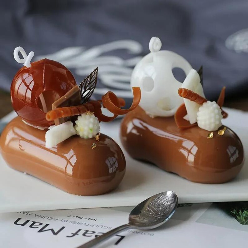 May include: Two chocolate mousse desserts decorated with caramel spheres, white chocolate, and gold leaf. The desserts are on a white plate.