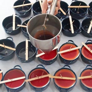 Puede incluir: Una persona vierte cera roja en peque&ntilde;os cuencos de cer&aacute;mica azul con palitos de madera y mechas. Los cuencos est&aacute;n dispuestos en filas sobre una superficie blanca.