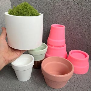 Könnte beinhalten: Eine Sammlung verschieden großer, leerer Blumenkübel aus Keramik und Silikon in den Farben Weiß, Rosa und Pfirsich. Ein weißer Topf enthält grünes Moos. Die Töpfe sind zylindrisch, konisch und abgerundet.