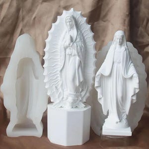 Może przedstawiać: Trzy białe religijne posągi Matki Boskiej. Jeden to forma, a dwa pozostałe to gotowe posągi. Posągi są szczegółowe z płynącymi szatami i aureolami. Centralny posąg stoi na białej ośmiokątnej podstawie.