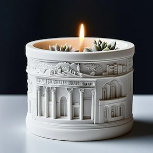 Puede incluir: Un portavelas cilíndrico blanco con un relieve detallado de arquitectura clásica. El portavelas presenta una vela encendida y pequeñas suculentas. El diseño incluye columnas, edificios y figuras, creando una estética clásica.