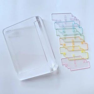 Puede incluir: Una caja rectangular de acrílico transparente y una pila de etiquetas de colores translúcidos. Las etiquetas son en tonos de rosa, amarillo, naranja, verde y azul, cada una con texto japonés. La caja está sobre una superficie blanca.