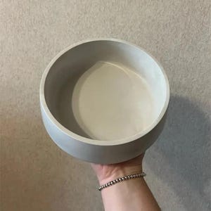 Könnte beinhalten: Eine hellgraue, runde Schüssel mit glatter Innen- und Außenseite. Die Schüssel wird von einer Person gehalten. Das Design der Schüssel ist schlicht und modern, für verschiedene Zwecke geeignet. Die Farbe der Schüssel ist ein neutraler Ton.