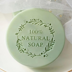 Puede incluir: Jabón redondo verde claro con las palabras "100% NATURAL SOAP" en el centro, rodeado por un diseño decorativo de hojas y puntos. El jabón está sobre un plato blanco, con una tela blanca y transparente en el fondo.