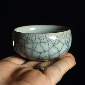 May include: A small, light grey ceramic teacup with a crackled glaze. The cup is round and has a slightly flared lip.