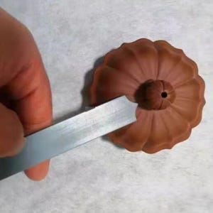 May include: A brown, flower-shaped ceramic piece with a central hole. A metal tool is held near the ceramic, possibly for shaping or detailing. The piece has a textured surface and a detailed petal design.
