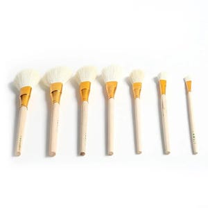 May include: A set of seven makeup brushes arranged in a row from largest to smallest. Each brush features soft white bristles, a shiny gold metal ferrule, and a light-colored wooden handle. The brushes vary in width and bristle shape for application.
