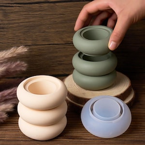 Op de afbeelding: Een verzameling kleine, ronde, containerachtige objecten in verschillende kleuren. Er zijn er drie gestapeld in lichtbeige, drie in gedempt groen en één in doorschijnend blauw. De objecten hebben een cirkelvormige opening aan de bovenkant en een afgeronde vorm.