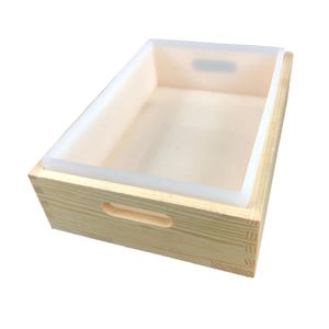 May include: A rectangular wooden soap mold with a light-colored interior and a white silicone liner. The mold has a natural wood grain finish and cut-out handles on the sides. The silicone liner sits inside the wooden frame.