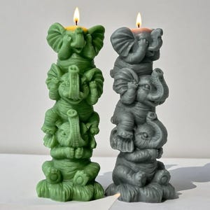 以下が含まれることがあります： 緑とグレーの2つの象の形をしたキャンドル。それぞれ3つの象が積み重なっています。キャンドルには火が灯っており、黄色い炎が出ています。象は「見ざる、言わざる、聞かざる」のポーズをとっています。