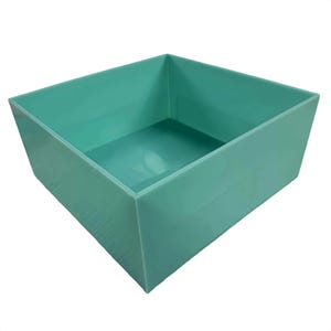 May include: A square, open-top storage bin in a solid, smooth, light teal colour. The container features straight, vertical walls and a flat base, presenting a clean, uniform appearance with no additional patterns, textures, or markings visible.