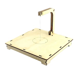May include: A wooden string art machine with a square base and a curved arm. The arm is attached to the base with a metal screw. A string is attached to the arm and is threaded through a hole in the center of the base.