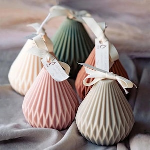 Puede incluir: Un conjunto de velas en forma de pera en varios colores pastel, incluyendo rosa, beige y verde. Cada vela tiene un diseño geométrico y está adornada con una cinta color crema y una pequeña etiqueta. Las velas están dispuestas sobre una tela suave de tono neutro.