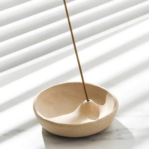 May include: A beige ceramic incense holder with a curved design and a small, raised area to hold the incense stick. The incense stick is brown and is placed in the holder. The holder is on a white surface.