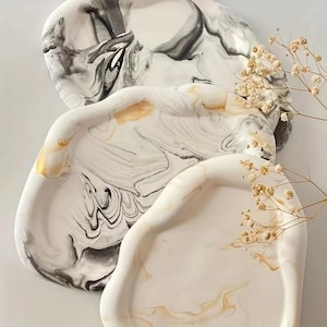 Puede incluir: Tres bandejas decorativas con diseño marmolado. Las bandejas son en tonos blanco, negro y dorado. Las bandejas tienen una forma orgánica y están dispuestas en una pila vertical. Una ramita de flores secas está en el fondo.
