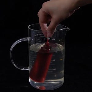 May include: A red candle is being lowered into a clear glass measuring cup filled with water. The measuring cup has markings in milliliters.