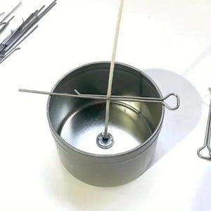 May include: A silver metal candle tin with a wick centered inside. A metal wick centering tool is placed across the top of the tin. Additional metal wick centering tools and wicks are visible in the background. Candle making supplies.