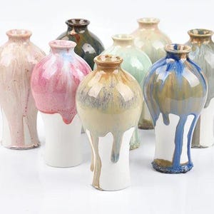Può includere: Una collezione di vasi in ceramica fatti a mano in vari colori, tra cui rosa, blu, verde e marrone. Ogni vaso ha un design di smalto unico, con alcuni che presentano un effetto gocciolante. I vasi hanno una base bianca e un collo stretto.
