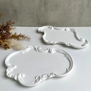 May include: Two white decorative trays with ornate, scroll-like edges. The trays have a classic, vintage design and are placed on a white surface. The trays are empty and ready for use.