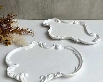 May include: Two white decorative trays with ornate, scroll-like edges. The trays have a classic, vintage design and are placed on a white surface. The trays are empty and ready for use.