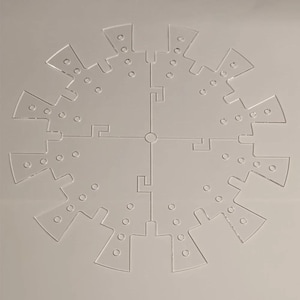 May include: A clear, circular, laser-cut acrylic piece. The design features a central circle with four intersecting lines, and a series of radiating, notched segments. Each segment has small, evenly spaced holes. The piece is designed to interlock.