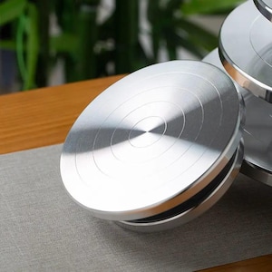 May include: A circular silver-colored metal cake turntable with a smooth, brushed finish and concentric circular grooves etched into the top surface. The rotating stand features a sturdy base and a flat, reflective top platform for display.