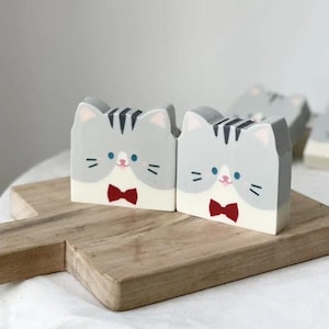 May include: Two gray cat-shaped decorative blocks with red bow ties and blue eyes sit on a wooden cutting board. The blocks have black stripes on their heads and white faces. A third block is visible in the background.