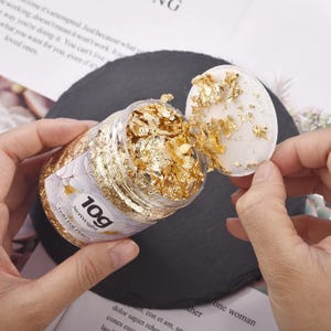 Peut inclure: Un pot transparent rempli de flocons de feuilles d'or, avec le couvercle partiellement retiré. L'étiquette indique "10g NetWeight Gold Foil Flakes". Le pot est tenu par les mains d'une personne, avec le couvercle dans l'autre main. Le fond est une surface gris foncé.
