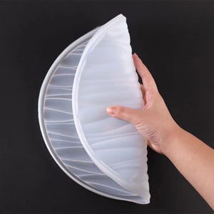 May include: A translucent white silicone mold, possibly for baking or crafting, is held open, revealing its interior. The mold has a wavy, textured surface and a rounded shape. The background is a solid black.