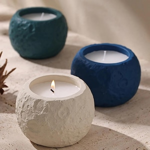 May include: Three spherical candles in shades of blue and off-white. The front candle is lit, showing a small flame. The candle holders have a textured, stone-like appearance. The candles are a decorative home accent.