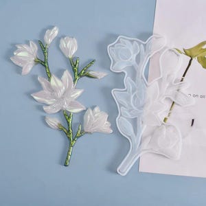 May include: A decorative floral branch ornament with iridescent white petals and a metallic green stem alongside a matching translucent silicone mold. The ornament features multiple blooming flowers and buds arranged on a single branch structure.
