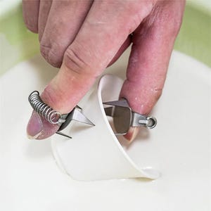 May include: A close-up of a finger wearing a spring-loaded ring with a small, sharp blade attached. The blade is being used to cut a piece of white material.