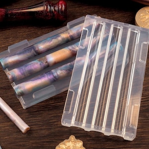 May include: A clear silicone mold with multiple slots, holding several colorful pens with a marbled design in shades of purple, blue, and gold. The pens are partially visible, with a matching pen lying separately. The background is a dark wood surface.