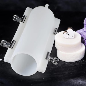 Pode incluir: Um molde de silicone cilíndrico branco translúcido fixado com quatro clipes de metal prateados, posicionado ao lado de dois sabonetes redondos brancos empilhados com um pequeno ornamento de madeira em forma de coração com uma estrela.