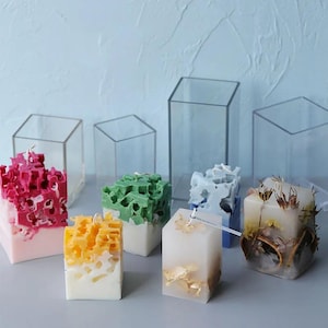 Peut inclure: Assortiment de bougies artisanales de différentes couleurs et motifs. Les bougies sont de forme carrée, certaines avec des motifs ajourés texturés dans des tons de rose, vert, jaune et bleu. Des moules carrés transparents entourent certaines bougies.
