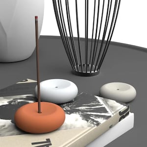 May include: An orange incense holder with a brown incense stick, two white incense holders, and a black metal decorative piece. The items are on a book with text and a grey surface.